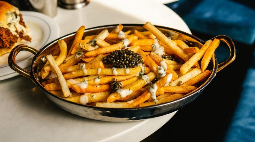 Golden fries topped with caviar and a creamy white sauce in a metal bowl with gold handles.