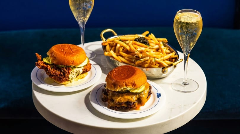 Two burgers, a basket of fries with caviar, and two champagne flutes on a white table.