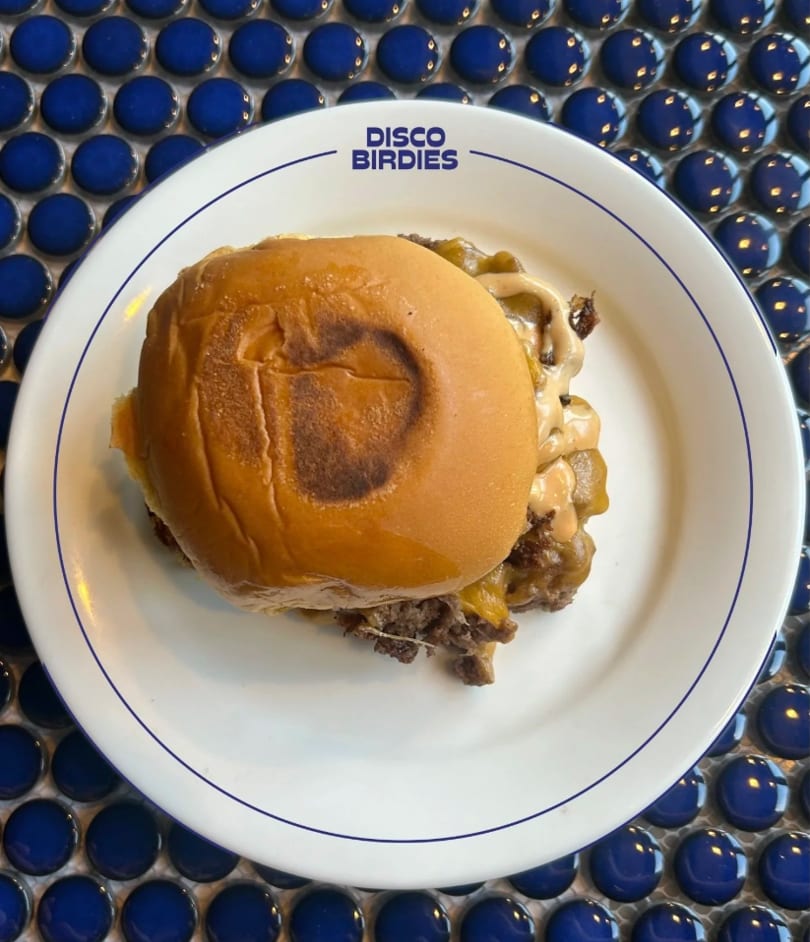 A Disco Birdies burger with melted cheese and sauce on a white plate over blue tiles.