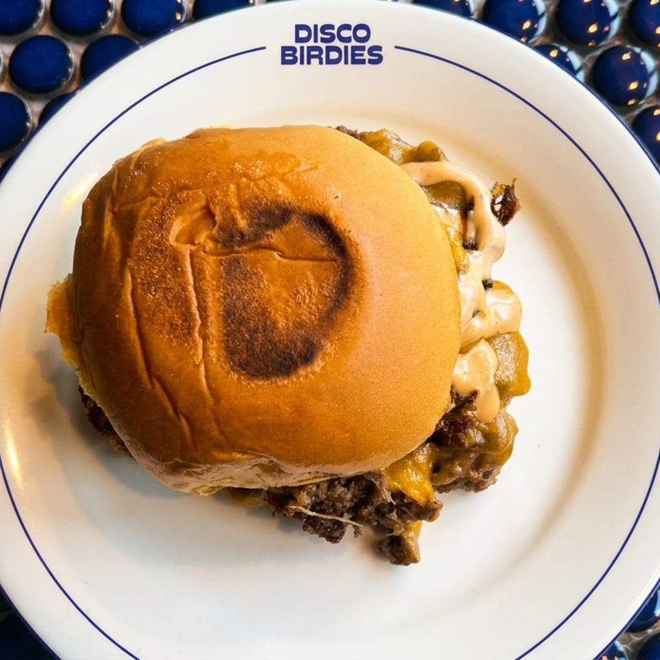 A Disco Birdies smash burger with melted cheese and sauce on a white plate with blue trim.