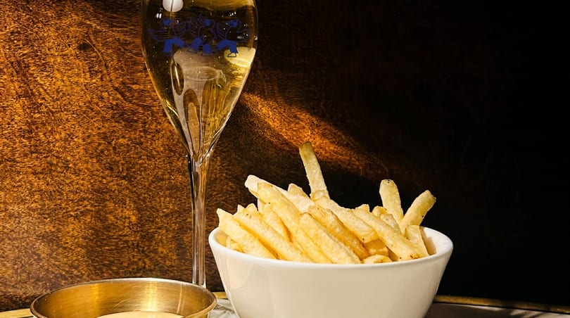 A flute of champagne with a blue logo next to a white bowl of french fries and a dipping sauce.